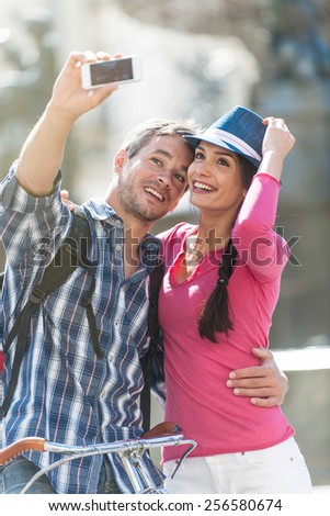 A couple is taking a selfie on their smartphone in the city center. The woman is wearing a blue hat. The grey hair man is wearing a backpack. They are looking at the camera with expressive faces.