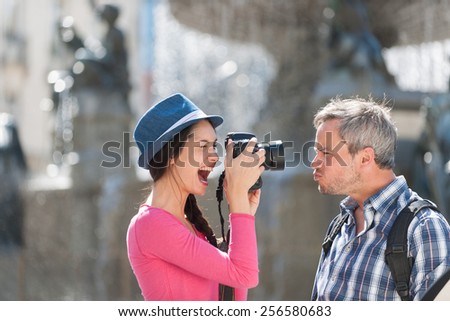 A loving couple is taking photos in the city center. The woman is taking a closed picture of the grey hair man with a beard and a black backpack while he does grimaces
