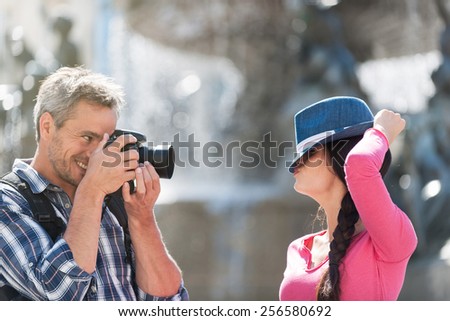 A couple is taking photos in the city center. The grey hair man with a beard and a black backpack is taking a closed picture of the smiling woman with a blue hat . She is hiding behind her hat.