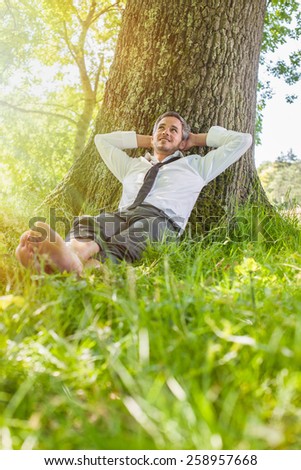 A nice looking man is sitting against a tree in the grass, looking like he is dreaming awake.  He is relaxing, enjoying the shadow of the tree in a sunny day.