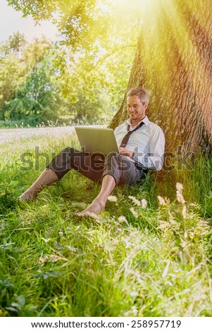 A nice looking grey hair man is sitting against a tree in the grass, looking at his computer. He is relaxing, enjoying the shadow of the tree in a sunny day.