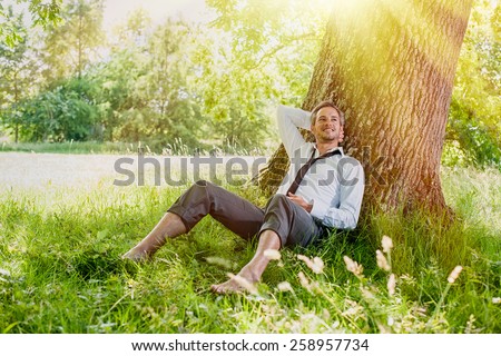 A nice looking man is sitting against a tree in the grass, looking like he is dreaming awake.  He is relaxing, enjoying the shadow of the tree in a sunny day.