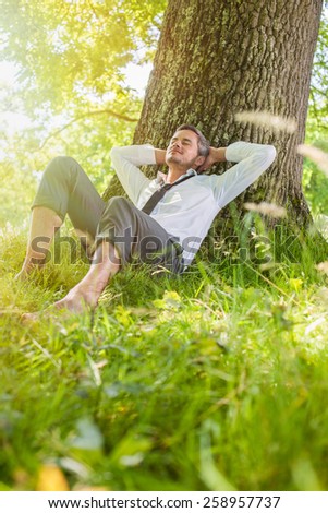 A nice looking man is sitting against a tree in the grass, looking like he is dreaming. He is relaxing, enjoying the shadow of the tree in a sunny day.