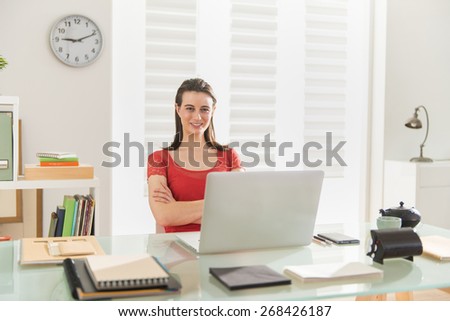 Looking at camera, sitting in her office, a modern businesswoman works on a project on her laptop, the office is modern and bright