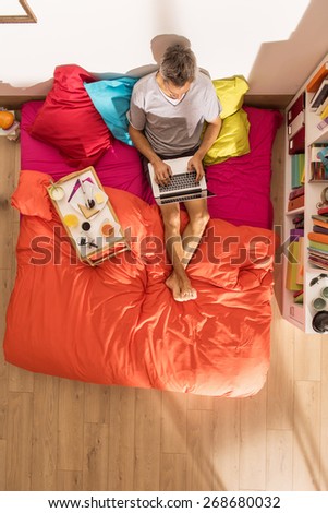 top view, single man using a laptop in bed by a sunny morning, his breakfast on the bed beside him, the bedding has bright colors, the flat is modern and luminous
