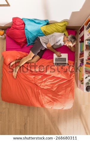From above, handsome man lying on bed using a laptop by a sunny morning, the bedding has bright colors. The flat is luminous and modern