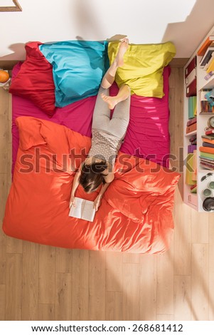 From above, young woman lying on bed reading a book, by a sunny morning, the bedding has bright colors. The flat is luminous and modern