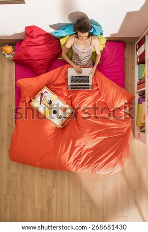 top view, single young woman using a laptop in bed by a sunny morning, her breakfast on the bed beside her, the bedding has bright colors