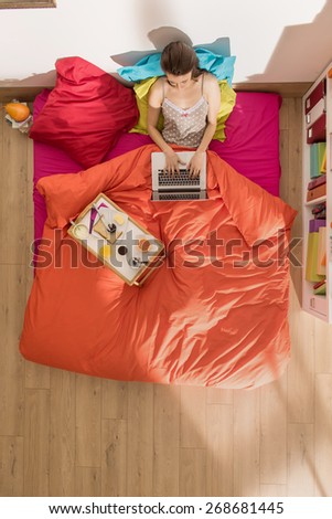 top view, single young woman using a laptop in bed by a sunny morning, her breakfast on the bed beside her, the bedding has bright colors