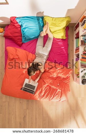 From above, young woman lying on bed using a laptop by a sunny morning, the bedding has bright colors. The flat is luminous and modern
