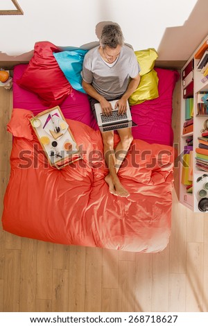top view, single man using a laptop in bed by a sunny morning, his breakfast on the bed beside him, the bedding has bright colors, the flat is modern and luminous
