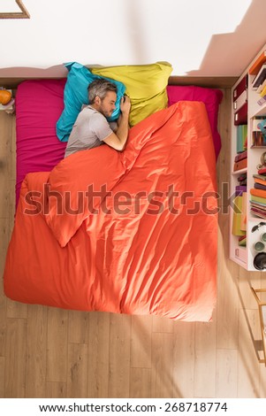 top view of a man lying in bed at bright colors, it has gray hair, a library is next to his bed in his sunny apartment
