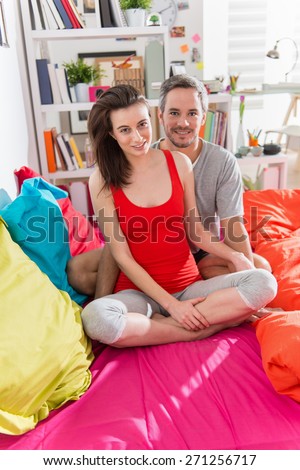 Looking at camera, a glamour couple in pajamas, sitting in bright colors\'s bed, man has gray hair, their apartment is modern and bright