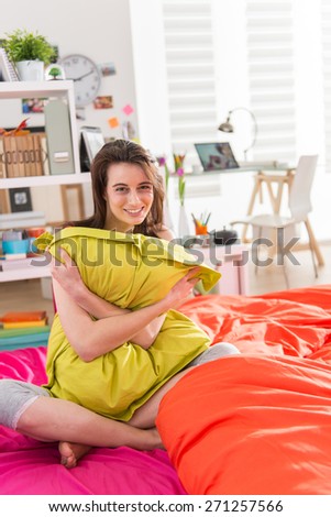 Looking at camera, a gorgeous young woman sitting in bed, clutching a green pillow in her arms,bedding is bright color, her sunny apartment is modern and bright