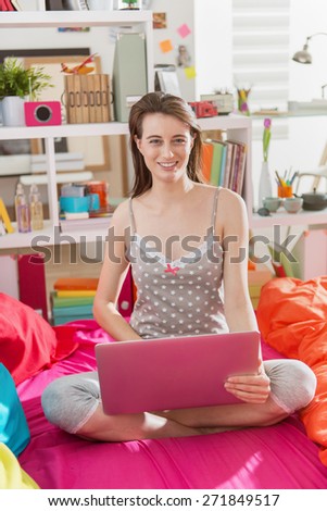 Looking at camera, a beautiful young woman in nightwear, sitting cross-legged in bed while surfing a laptop,bright color's bed and her interior is bright and contemporary