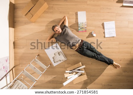 view from above, looking at the camera a modern man lying on the floor of his new home, there are blueprints and samples colors to help him to think about his new interior decoration
