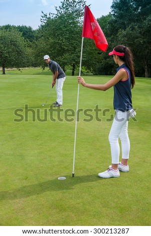 Nice couple playing golf on a green course during a sunny day. The woman is holding the red flag pole while her partner is swinging his club, aiming at the hole They are wearing sportswear outfits