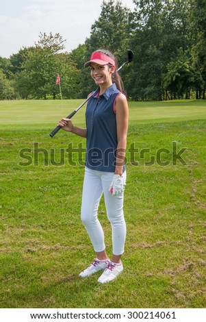 Portrait of a young woman holding a club on her shoulder on a golf course. She is standing, smiling at camera wearing a red cap and a sportswear outfit