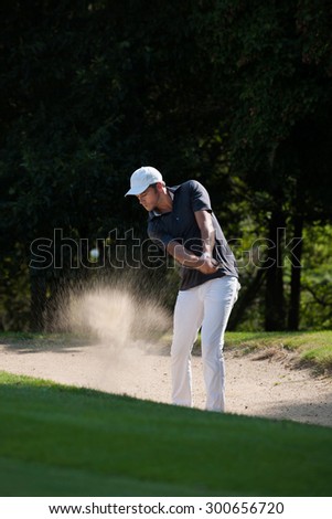 Portrait of a middle aged man trying to shoot his ball out of the bunker. He is playing golf with a white cap and the sand is flying around him