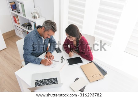Top view of a couple of architects working on ideas. A grey hair man and a woman are sitting at a white table in an open space wearing casual clothes, they are drawing plans on a spiral notebook