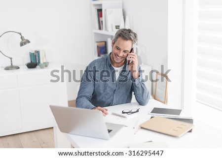 Portrait of a nice smiling grey hair man with beard, he is sitting at a white table, talking on the phone with his laptop, tablet, notebook and glasses in front of him. Focus on the man