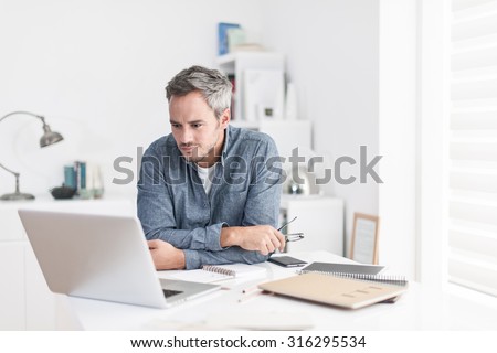 Portrait of a nice smiling grey hair man with beard holding his glasses, working at home on some project, he is sitting at a white table looking at his laptop in front of him. Focus on the man