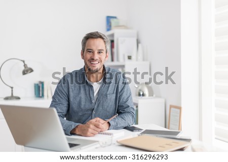Portrait of a nice smiling grey hair man with beard, working at home on some project, he is sitting at a white table looking at camera, writing ideas with his laptop in front of him. Focus on the man