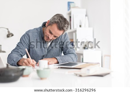 Portrait of a nice smiling grey hair man with beard, working at home on some project, he is sitting at a white table, writing creative ideas on papers with tea in front of him. Focus on the man