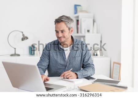 Portrait of a smiling grey hair man with beard, working at home on some project, he is sitting at a white table, drawing ideas on a white notebook, with his laptop in front of him. Focus on the man