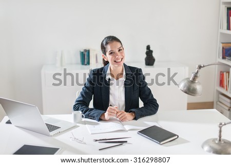 Close up of a smiling successful middle age businesswoman with dark hair sitting at her white desk in her office. She is wearing a black suit jacket, holding a pen, her laptop and coffee next to her