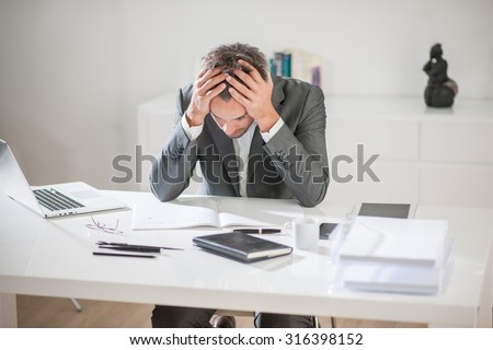 Portrait of a businessman with grey hair and beard having a hard time with some ideas. He is sitting at his white desk, holding his head in his hands above his notebook like if he was trying to think.