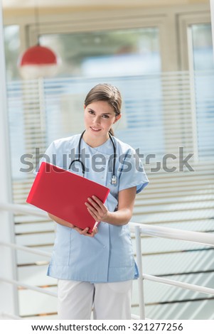 A middle aged nurse in blue uniform is looking at a red patient file. She has her stethoscope around her neck and a ponytail. She is standing in the hospital hallway and looking at camera.