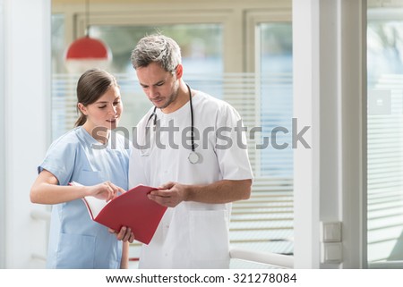 A nice grey hair doctor with beard and a nurse are examining a patient file He is wearing his white coat, his stethoscope around his neck. They are looking at camera in front of hospital room