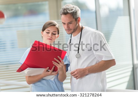 A nice grey hair doctor with beard and a nurse are examining a patient file He is wearing his white coat, his stethoscope around his neck. They are standing in front of hospital room with glass walls
