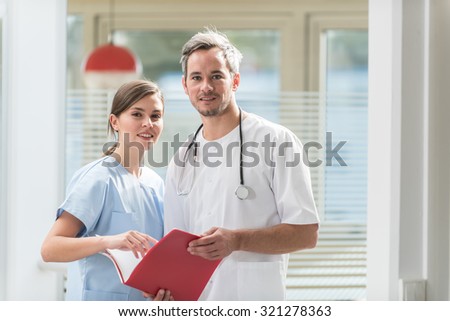 A nice grey hair doctor with beard and a nurse are examining a patient file He is wearing his white coat, his stethoscope around his neck. They are looking at camera in front of hospital room