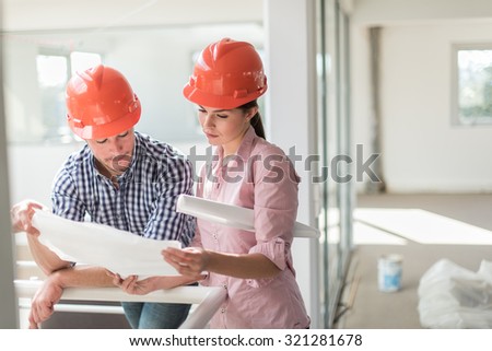 A female architect and a foreman examining blueprints on a construction site. they are standing in a luminous open space, leaning against a white guardrail. They are wearing orange hard hats