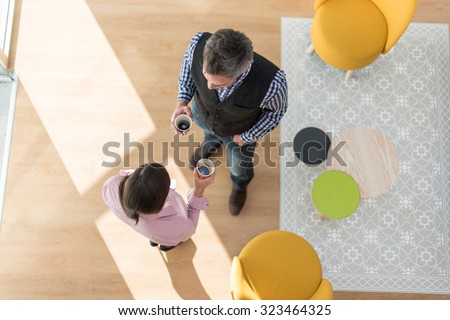 Top view of two co-workers having a break in a stylish vintage living room with wooden floor. The grey hair man and the woman are standing next to two orange seats, holding their coffee and talking