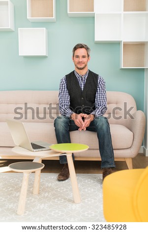 A grey hair man with beard and checkered blue shirt is sitting on a sofa in a stylish vintage waiting room with pastels colors. He is looking at camera, arms crossed, his laptop next to him