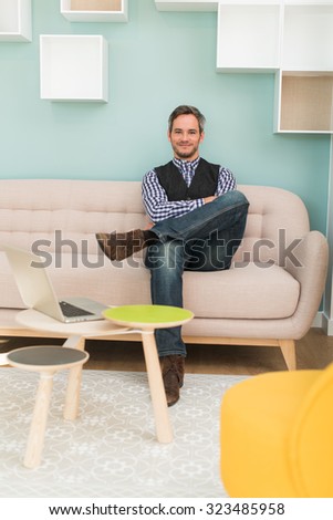 A grey hair man with beard and checkered blue shirt is sitting on a sofa in a stylish vintage open space with pastels colors. He is looking at camera, arms and legs crossed, his laptop next to him