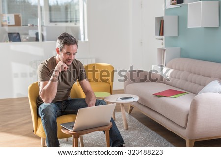 A middle aged grey hair man with beard in a stylish vintage living room with wooden floor. He is sitting in an orange chair with his laptop in front of him He is wearing casual clothes