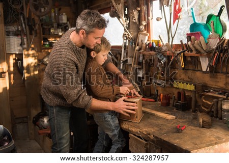 Father and his son working together in a wooden workshop. They are focused on building a wooden bird house, the son is knocking with a hammer while his grey hair father is holding the house