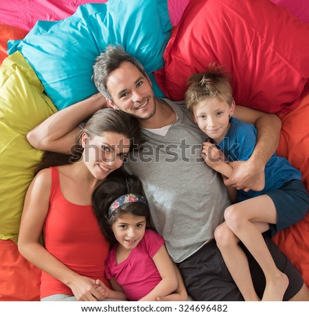 top view of a four people family laying on their sofa with large cushions. They are cuddling, the grey hair father is hugging his wife and his blond son and the mother is holding her daughter