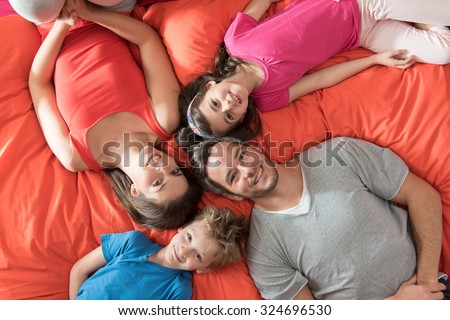 top view of a four people family laying on their big red bed with their heads next to each others. They are wearing colorful tops, smiling and looking at camera. The father has grey hair and a beard