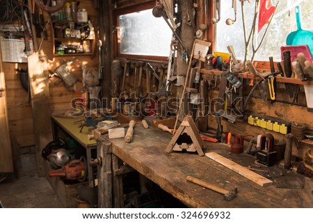 Picture of a rustic wooden workshop. Tools are everywhere, under and on the table, hanging from the ceiling. The last pieces of work on the table is a wooden birdhouse and a table leg