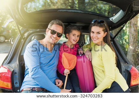 A smiling family looking at camera, they are sitting in the trunk of their car. They are leaving for the holidays. They are wearing colored pulls and holding a scoop net. The girl is seven years old