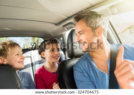 A grey hair father with beard is talking with his two kids in the car. The ten years old brother and sister are sitting in the back, they are wearing long sleeves shirts. The father have his seat belt