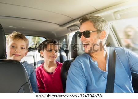 A grey hair father with beard and sunglasses is laughing with his two kids. They are in the car leaving for the weekend. The mother is in the background, looking at her phone.