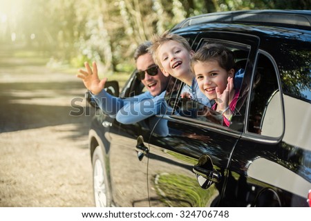 A father and his two kids are waving their hands through their car windows. they are on a country road. The brother and the sister are having fun, the grey hair man is wearing sunglasses