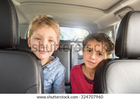 Two seven years old are sitting at the back of a car They are looking at camera,  stretching to reach the front seats The blond boy is wearing a blue shirt and the girl a pink top