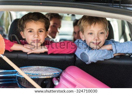 Rear view of a four people family in their car leaving for the holidays. The smiling little girl and boy are turned on their seat, looking at camera. A net and rackets are in the trunk